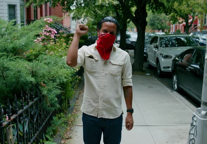 A man stands on a town sidewalk wearing a bandana face covering and clapping his hands