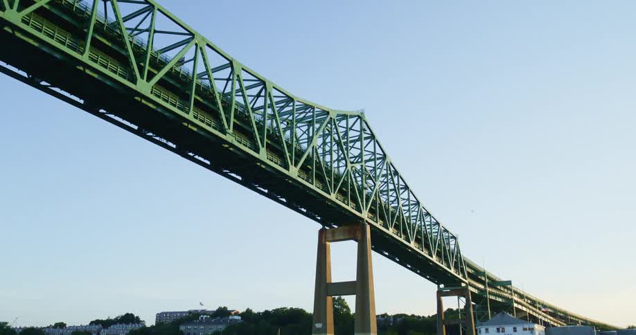 A long trestled bridge with many beams is stretching over a large body of water lined with buildings houses and boats