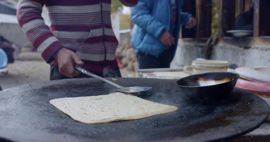 A man in a sweater flips a flat bread on an outdoor griddle then crosses his arms