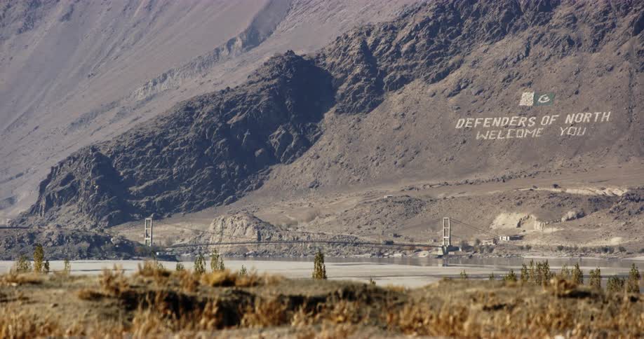 A bridge with vehicles over a river with plants in the foreground and a mountain in the background