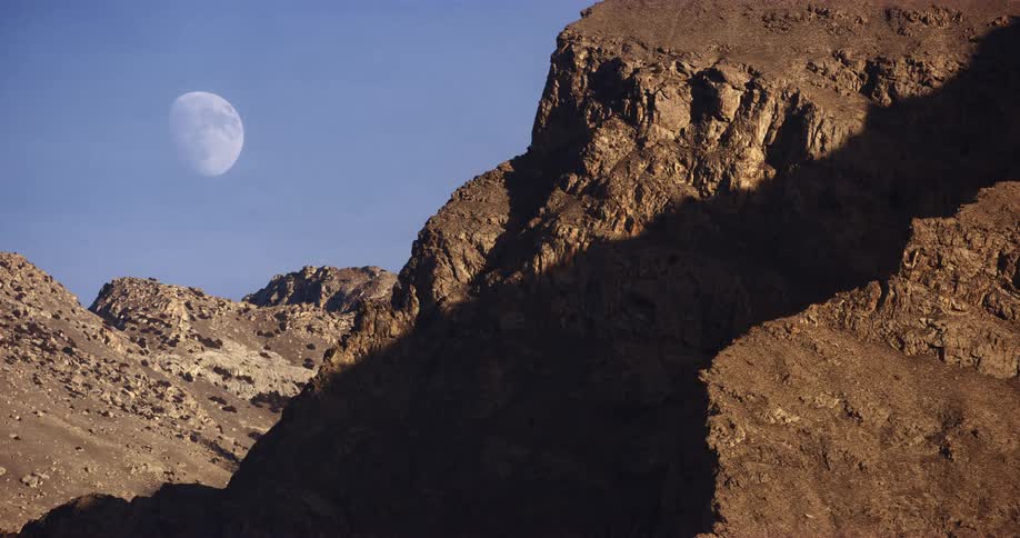 The waxing gibbous moon looms in the daytime sky over a brown barren mountain