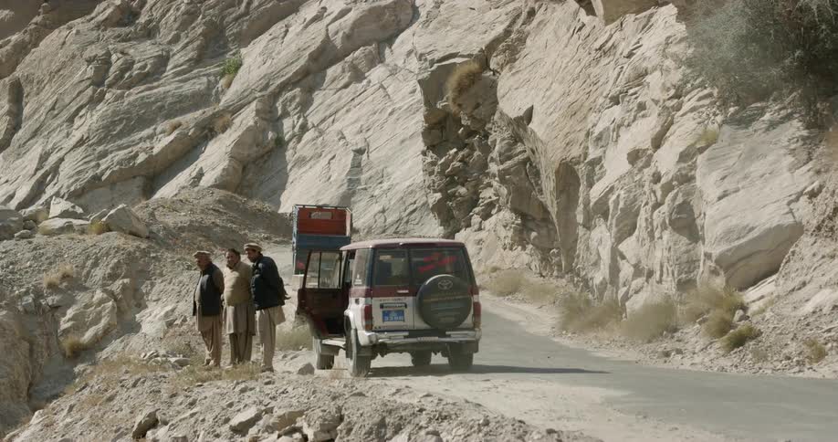 Four men stand next to their van and look over the edge of a cliff as vehicles drive by them