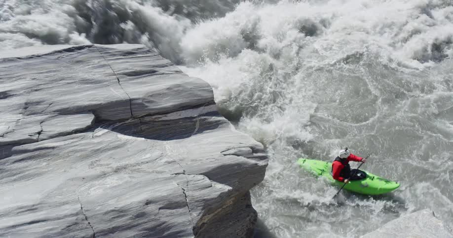 A person inside a green kayak on a rock pushes off into the raging rapid river below