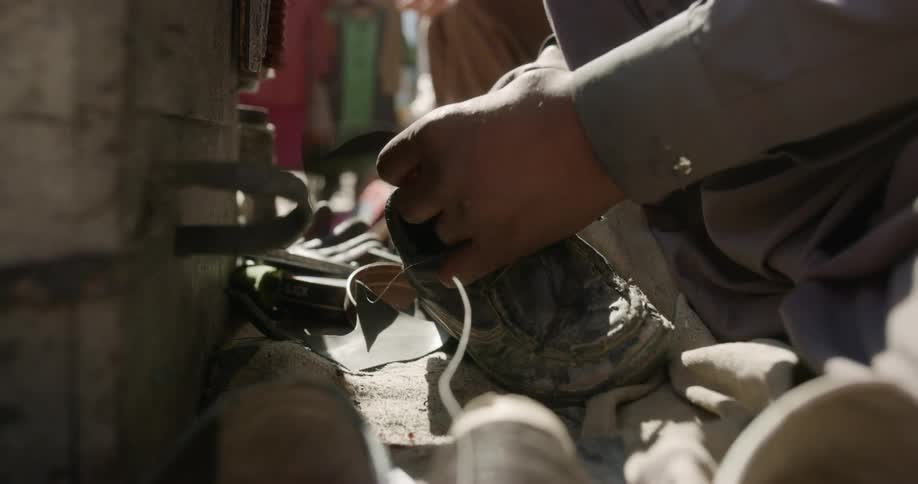 A kneeling man mends an old shoe with a needle and string