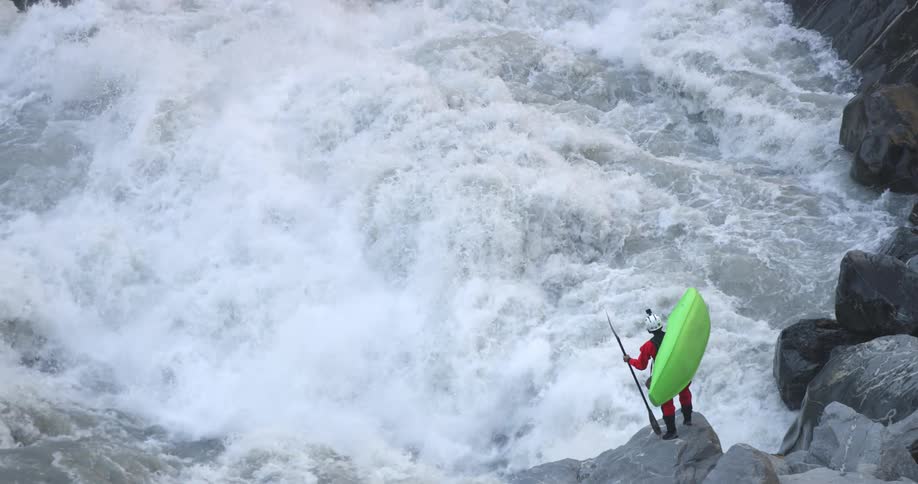 Man carrying a bright green kayak and a paddle looks at the turbulent rivers rapids from a rock