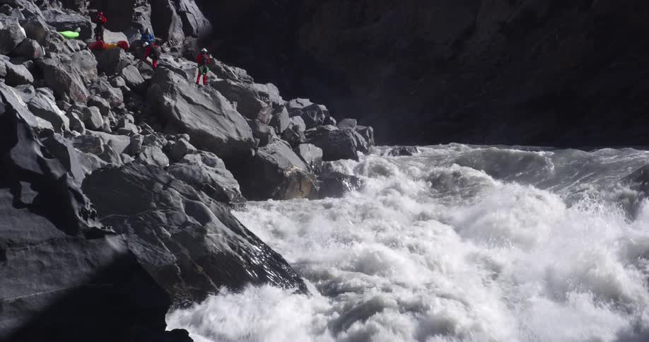 People wearing helmets stand on boulders near kayak looking down at rapids