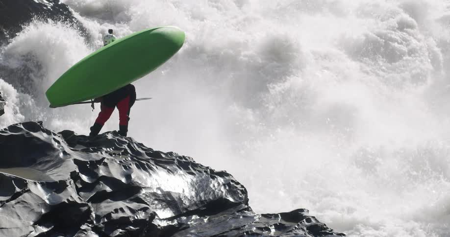 Person carrying a green kayak stands on the rocks looking at the turbulent waters of a rivers rapid