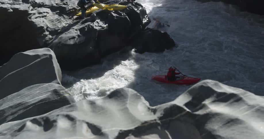 A pair of people sitting in their kayaks push themselves off a rock at the bottom of a canyon and into the violent river flowing through the canyons bottom