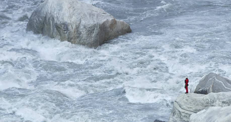 A person stands on a large rock on the edge of a river and watches the rapids