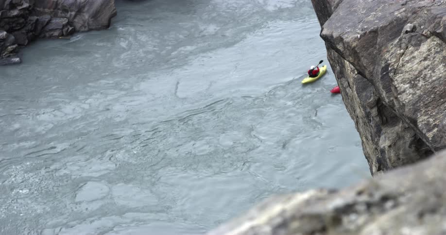 Two people in a yellow and red kayaks pedal slowly down a fast flowing river between mountains
