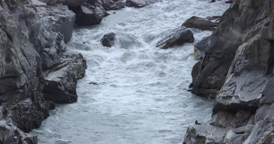 One person sits on a large rock and watches as another person kayaks through river rapids