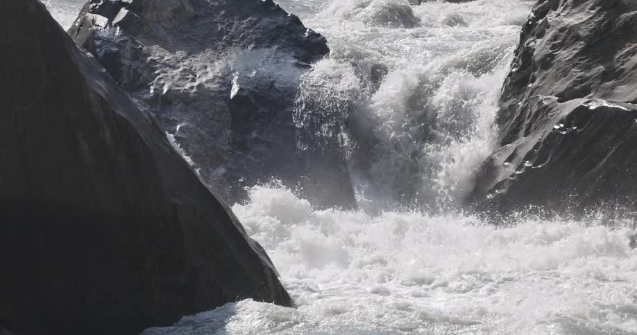 Water falls down low fall to churn into foam before continuing to flow past dark grey stone slabs