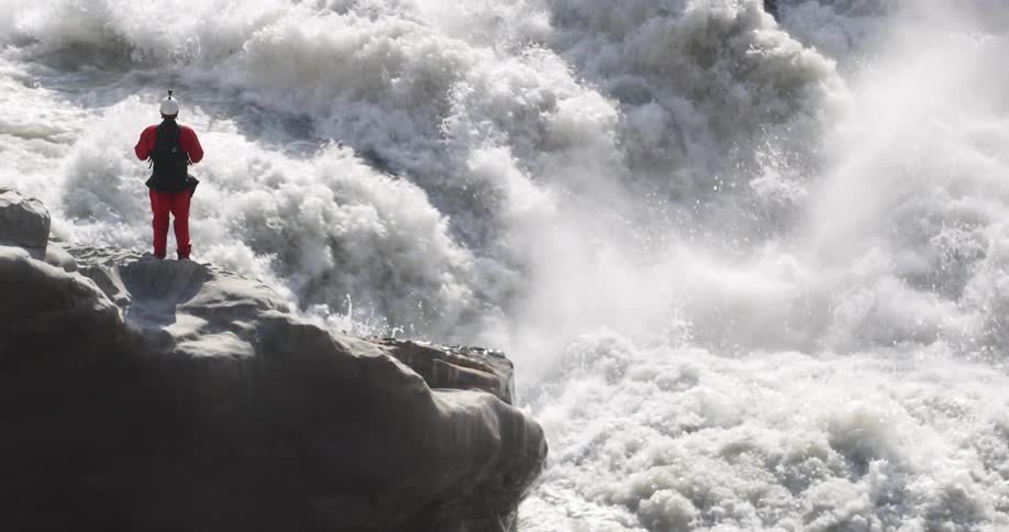 Hiker stands on a large rock while looking out at the crashing river rapids
