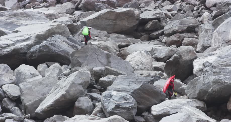 Two people carrying kayaks navigate through massive boulders on slope