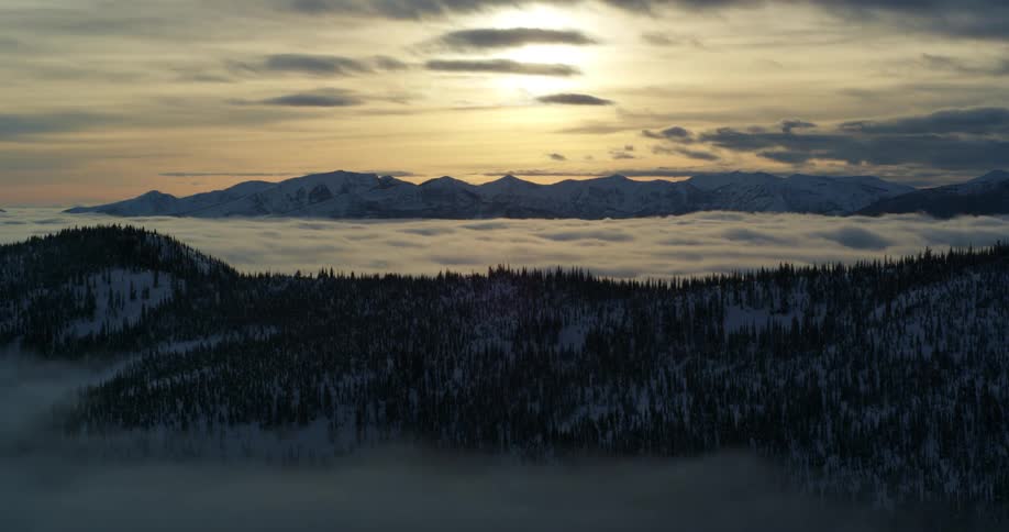 Clouds pass over a mountain ridge covered in a snow and evergreen forest during the day