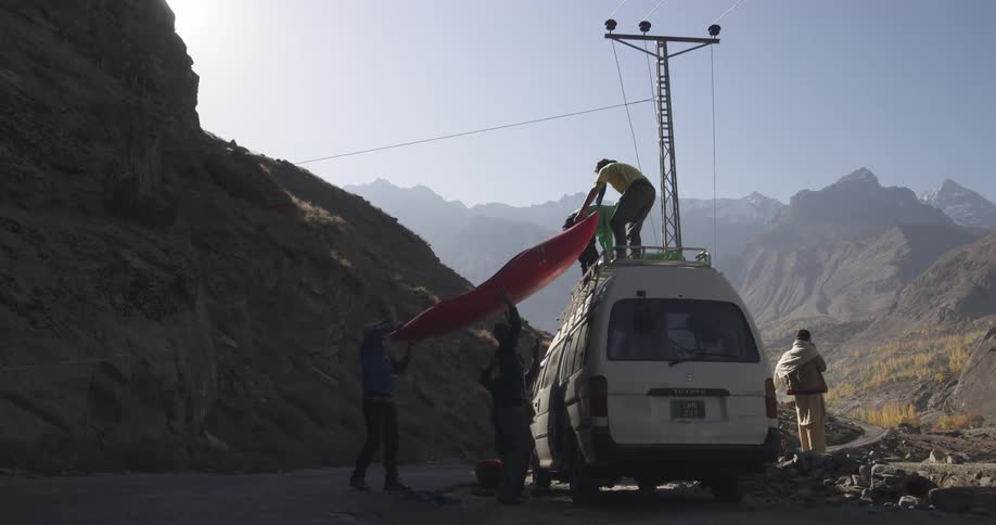 Two men standing on the roof of a van hand down colorful kayaks to the men below