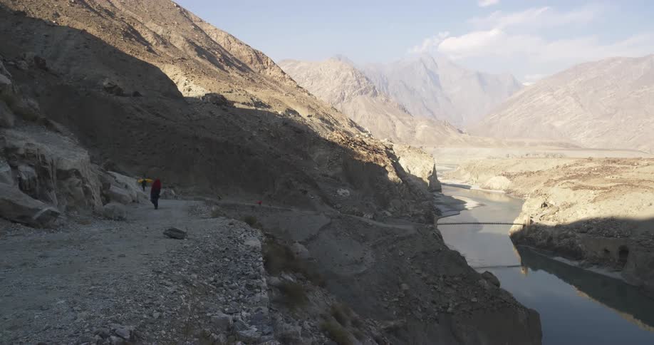 Two people carrying kayaks walk along the base of a mountain next to a river