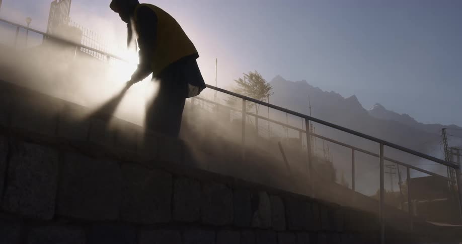 A person sweeping a sidewalk and raising dust with a mountain range and the sky in the background