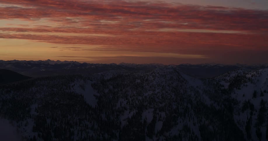 Clouds move in the colorful sky above snow covered mountains and trees at sunset