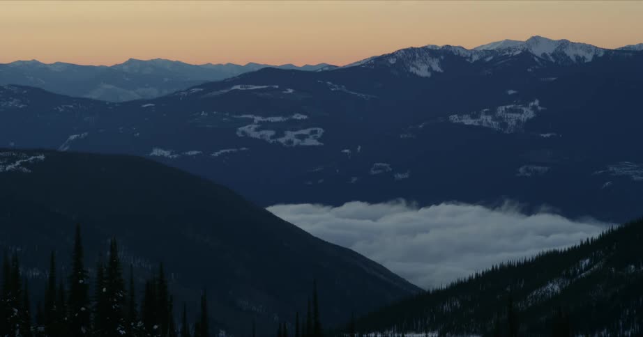 A mountain range with trees and snow with the sky and clouds in the background