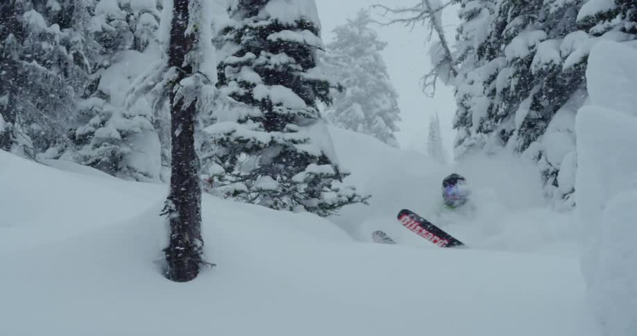 A skier flies off a hill on a slope and tries to stabilize themself as they land and continue on down the slope while heavy snow falls from the sky