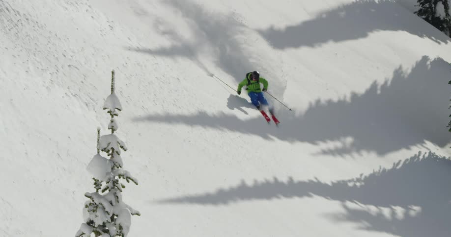 A person wearing a blue and green snow suit is skiing down a snowy slope flinging a trail of snow behind him