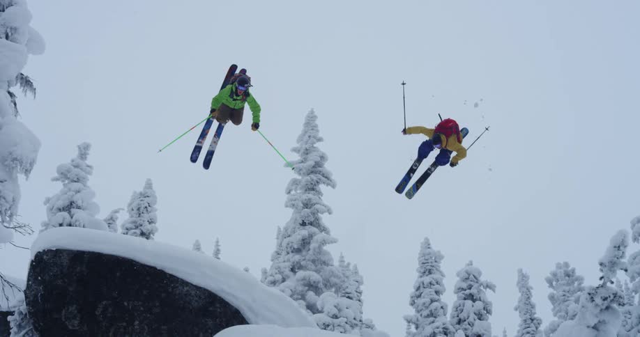 Two synchronized skiers jump over a rock into the snow covered slopes in the mountains