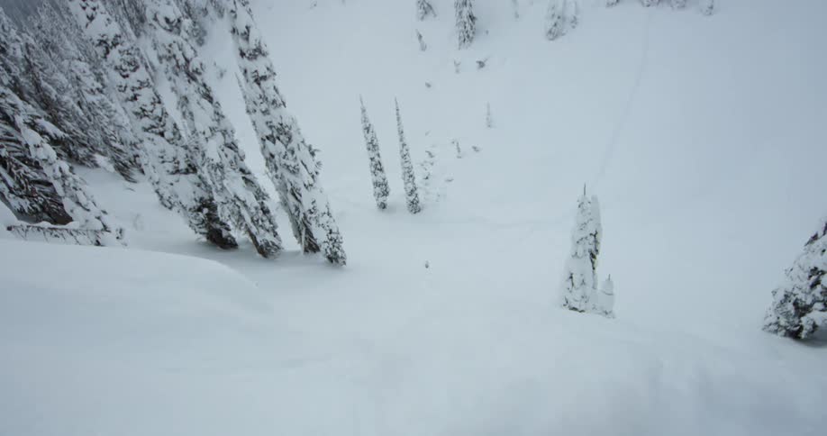 A snowboarder flies off a snowy cliff and spins in midair before landing on the mountain slope below and continuing downhill