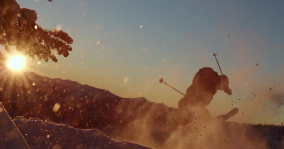 Skier jumps over a slope kicking snowflakes on the mountains at sunset
