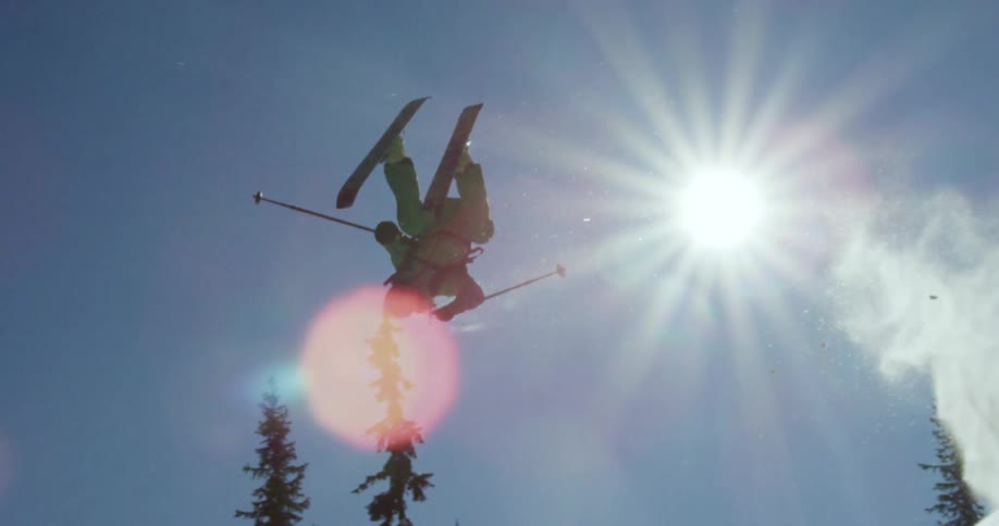 The sun shines down on a person flipping in the air as they ski down a mountain