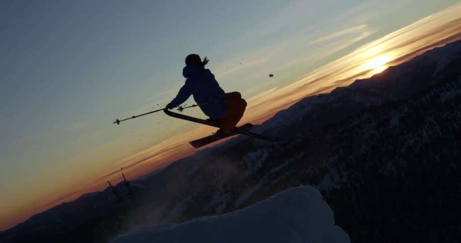 A person with a helmet and jacket skiing off a cliff with trees a mountain range and the sky in the background