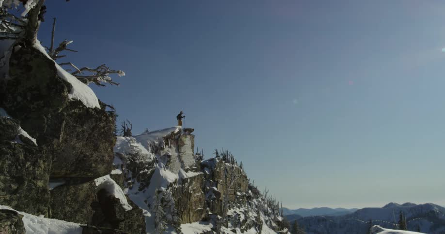 A man stands on a cliff in mountain range and looks over the landscape