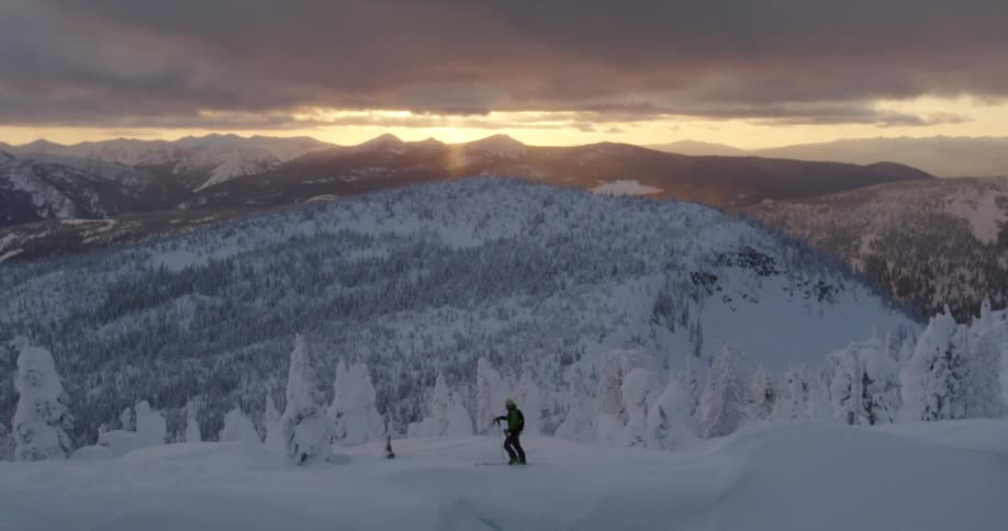 Person cross country skiing in snow covered forest in front of mountain range at sunset