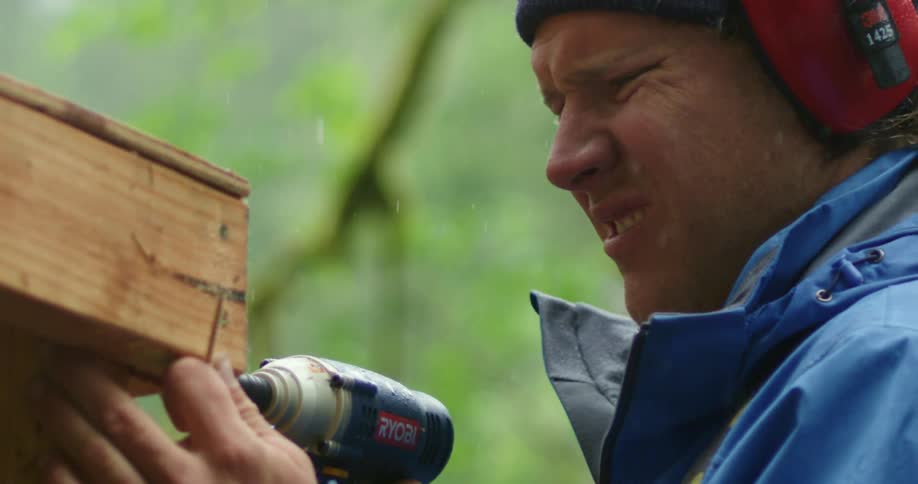 A man standing outside in the rain uses a drill on wood