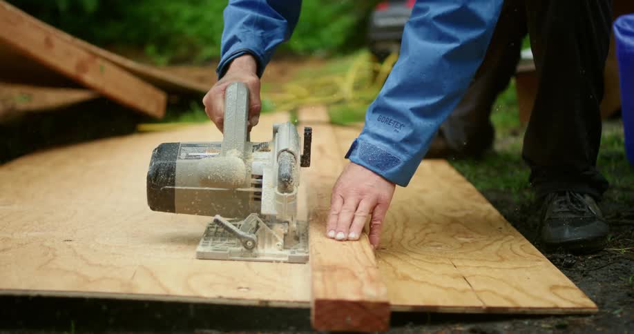 Man with power saw cuts plywood using two by four as a guide