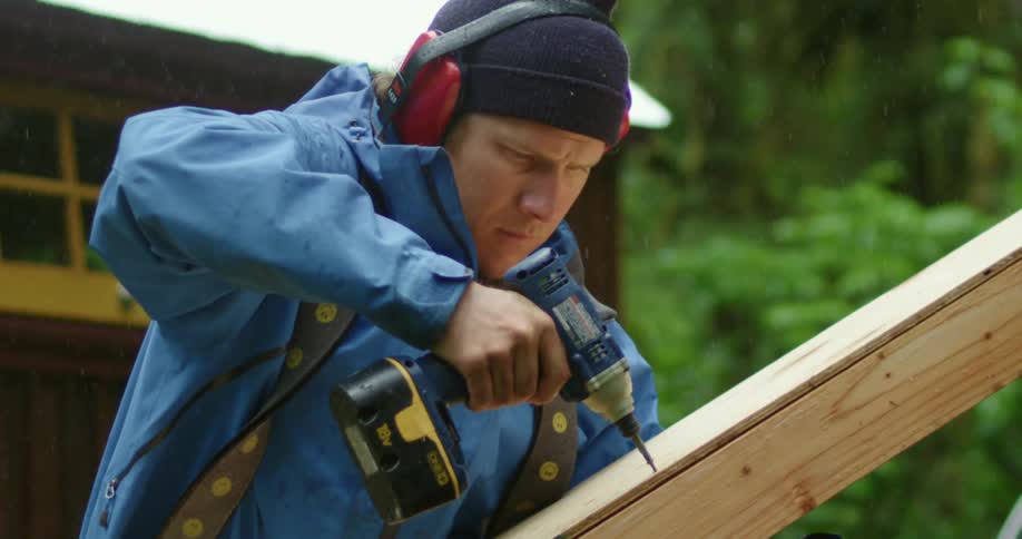 Carpenter drills a screw on a wooden beam outdoors under light rainfall