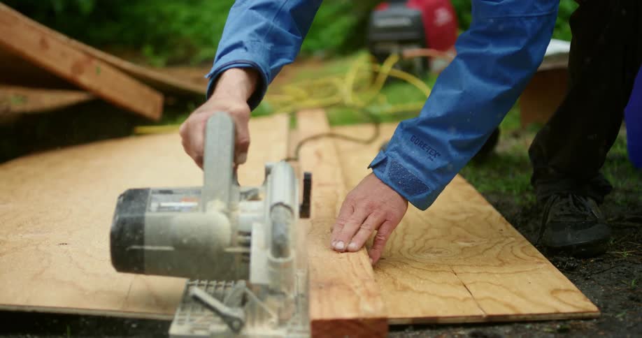 A kneeling man uses a circular saw to cut through wood on the ground