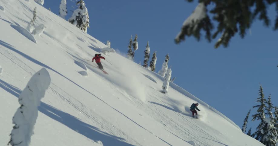 Two people ski down the snow covered slopes of a mountain under a clear blue sky