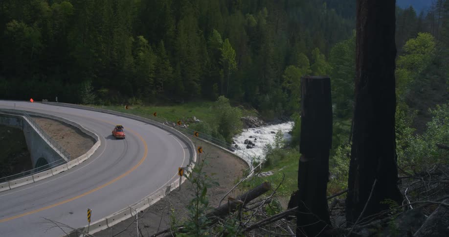 A vintage vehicle travels across a bridge at the base of a forested mountain