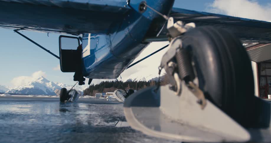 A pilot climbs into a small blue airplane parked at the edge of the water
