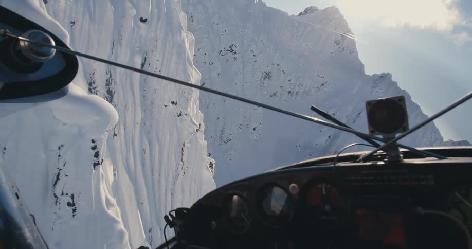 A person with a headset and sunglasses in an airplane with a mountain range and the sky in the background