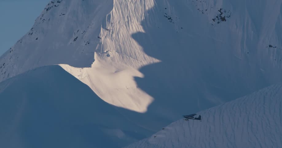 Small airplane flying over the snow covered peaks of a mountain range at sunset