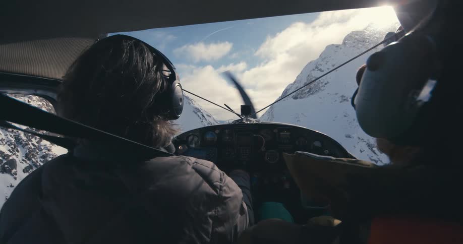 Two people sit in cockpit of aircraft flying between snow covered mountains