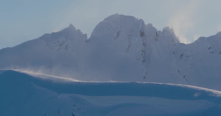 The wind blows up snow on a snow covered mountain during the day