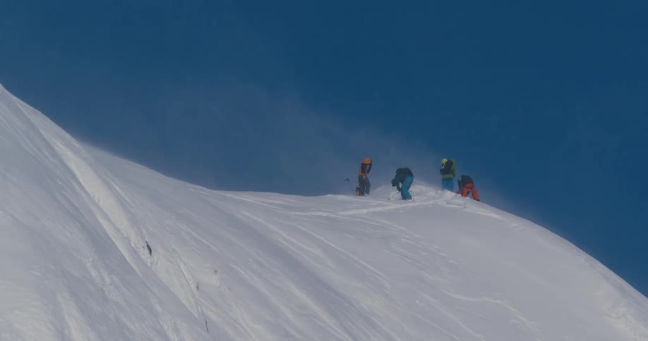 Wind blows over climbers in colorful winter gear atop of a snow covered mountain