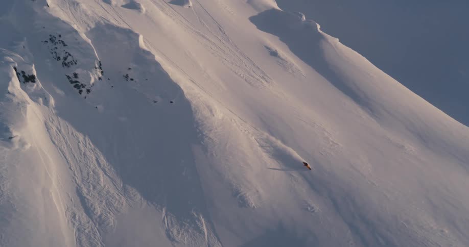 A snowboarder jumps down a steep snow covered slope then curves and rides through fresh powder