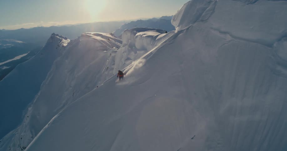 A snowboarder takes off from a mountain peak down a steep and narrow slope