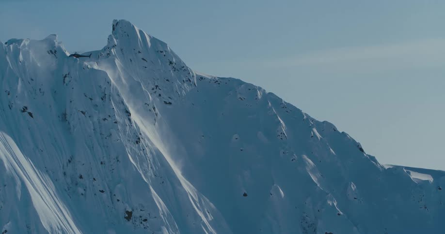 Helicopter hovers near top of mountain causing snow to drift down mountains steep slope under a lightly clouded sky
