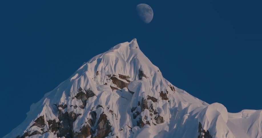 Time lapse of the moon passing over the snow covered peak of a mountain at sunset