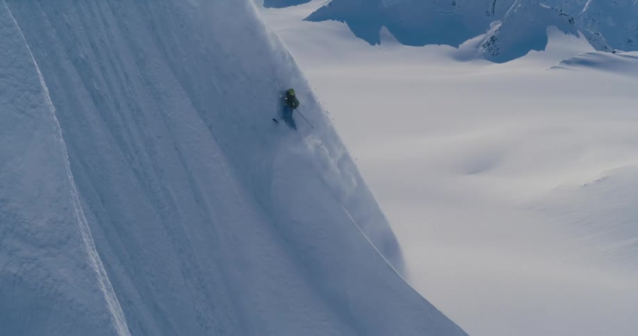 A person with a backpack skiing down a snowy slope with a mountain range in the background